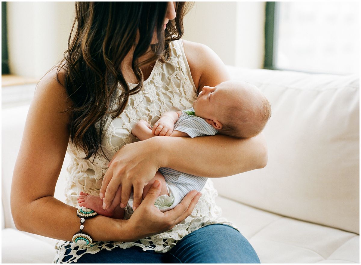 mom wearing a white crochet top and holding her newborn baby boy and sitting on white couch in NJ home