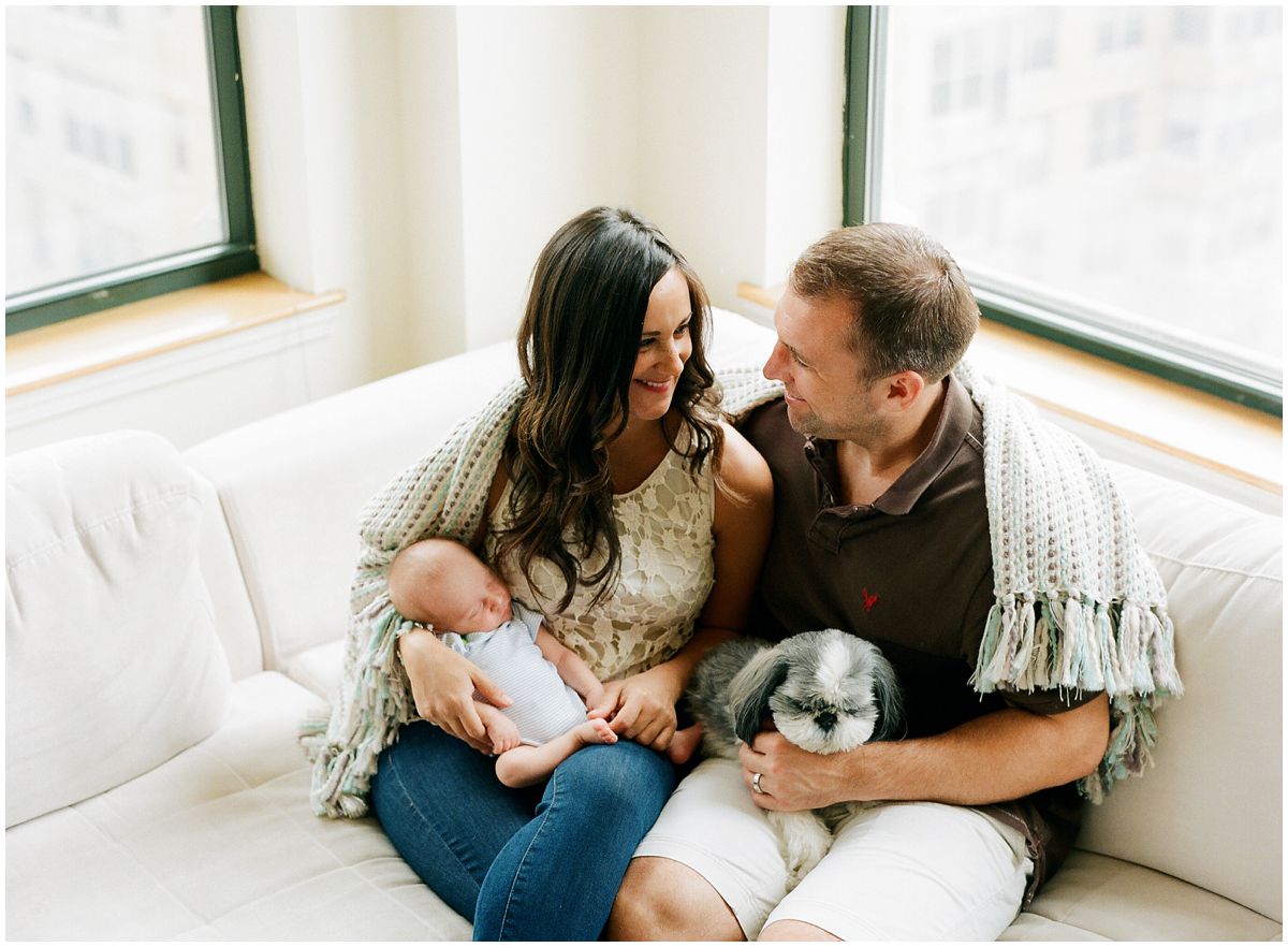 a couple is cozy in sharing a corner of a couch with their family dog and newborn son