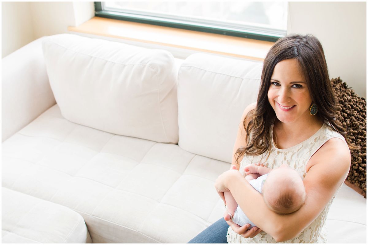 Newborn photographer captures smiling mom holding her newborn baby boy on a white couch by the window in Hoboken NJ
