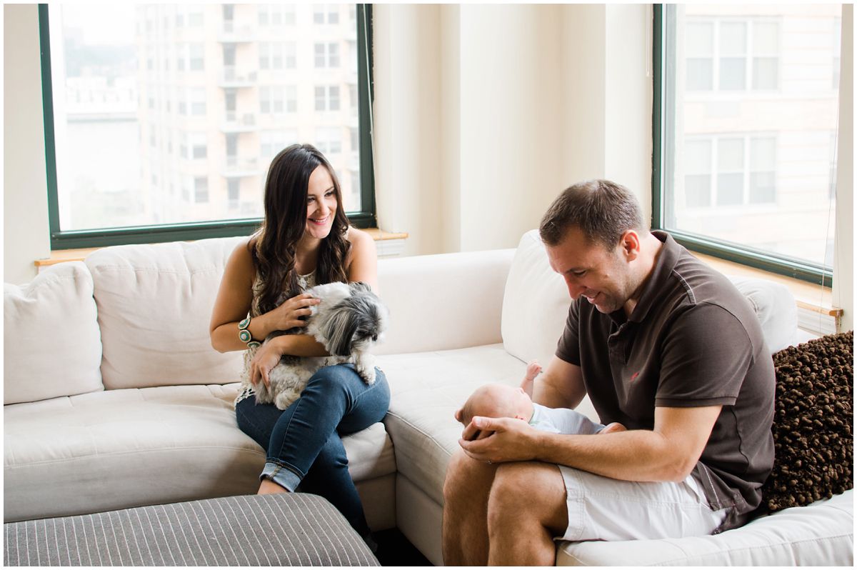 excited dad holding his newborn baby boy and smiling mom holding the furry dog on her lap in their Hoboken NJ home