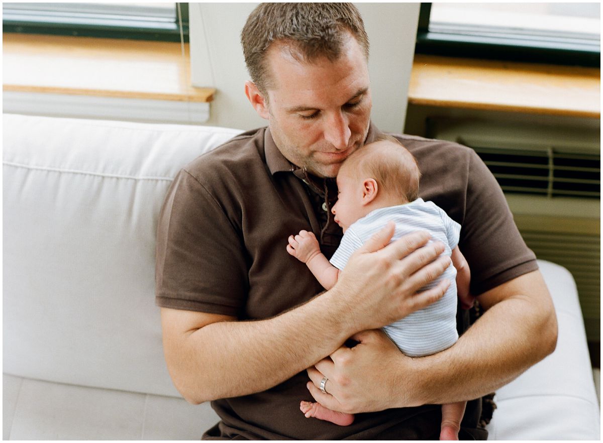 Newborn Photographer Hoboken NJ takes a photo of a dad snuggling with his baby boy on the white couch