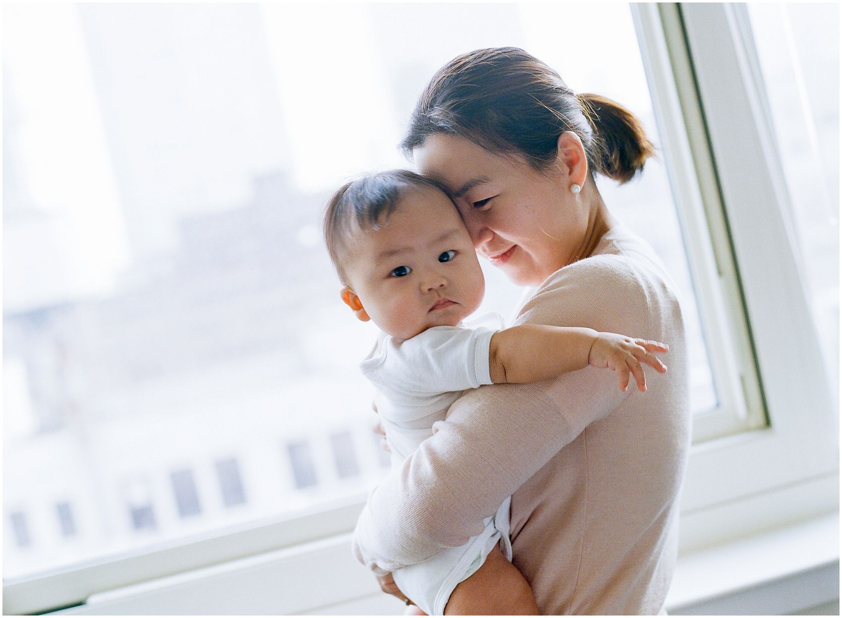 mom snuggling with her 6 months baby boy by the window overlooking NYC