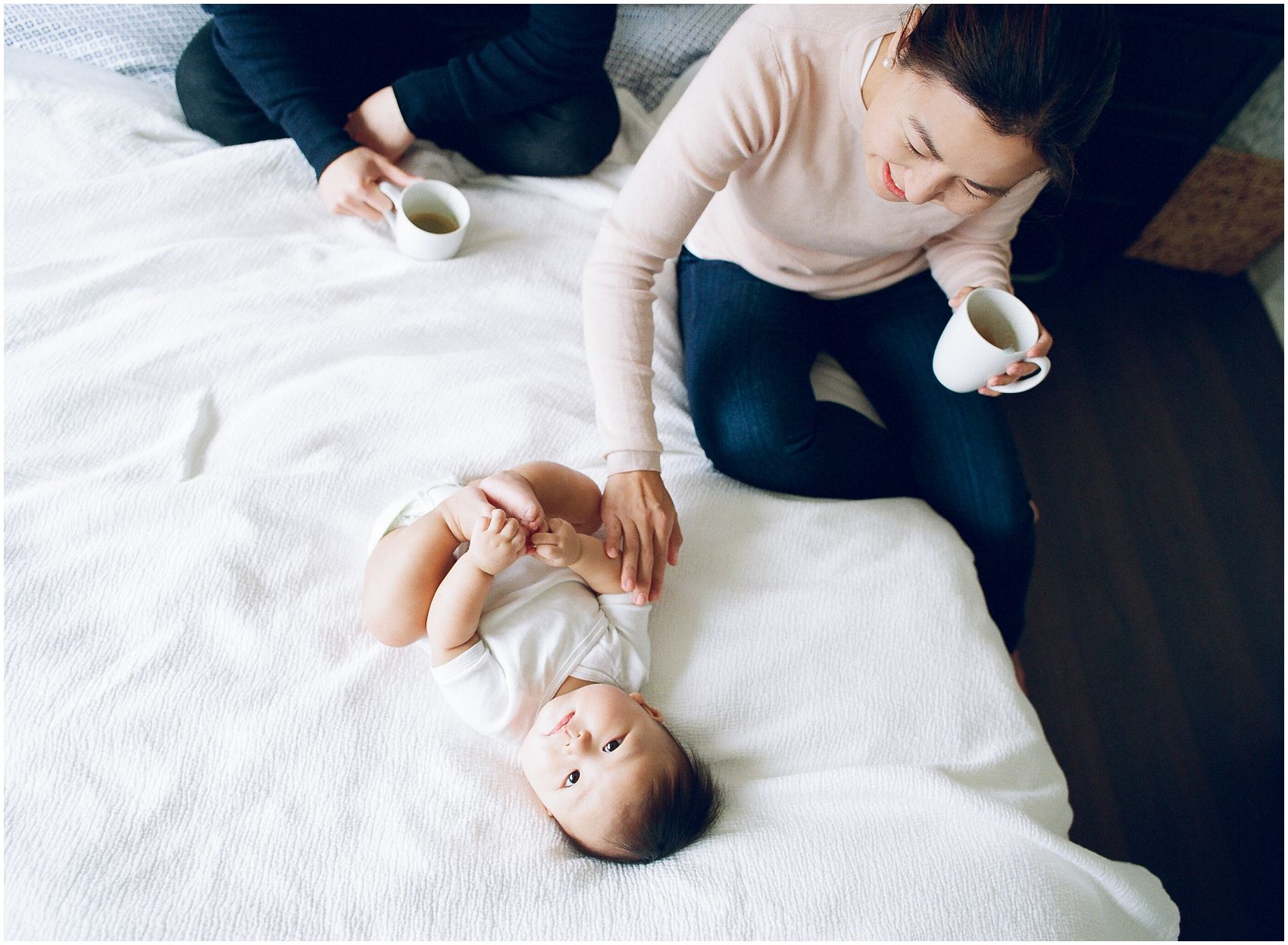 happy mom cuddling in white bed with a new baby and drinking coffee on a white bed in NJ