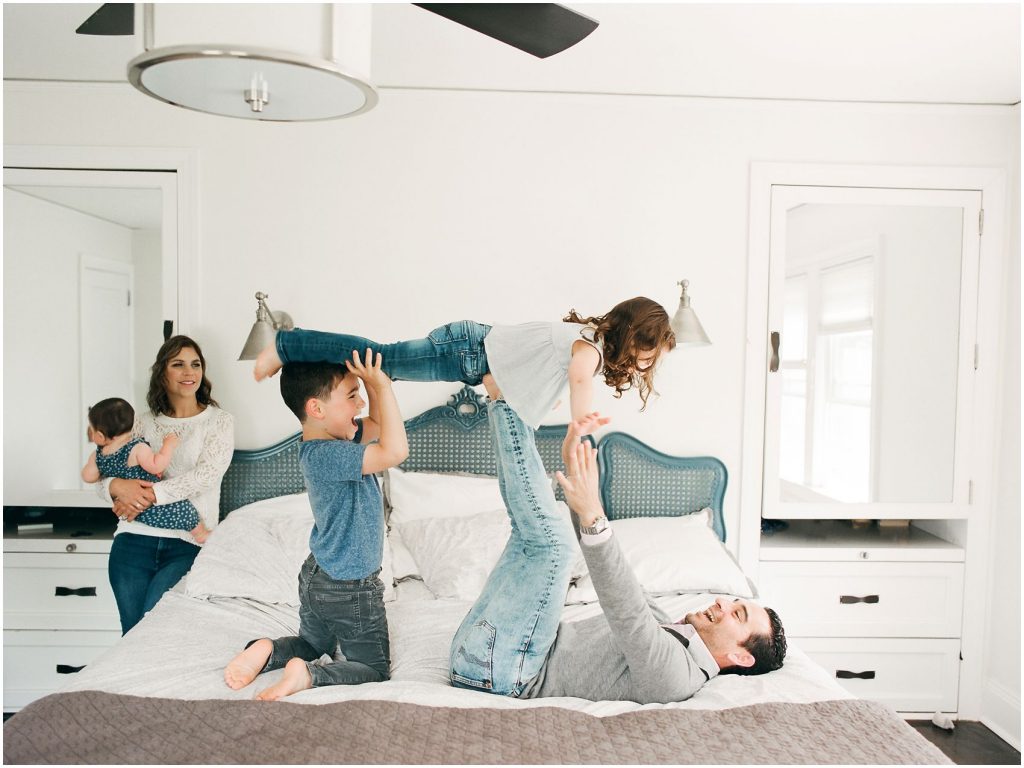 Father is playing airplane on bed with two kids while smiling mom looks on in family session in Montclair NJ