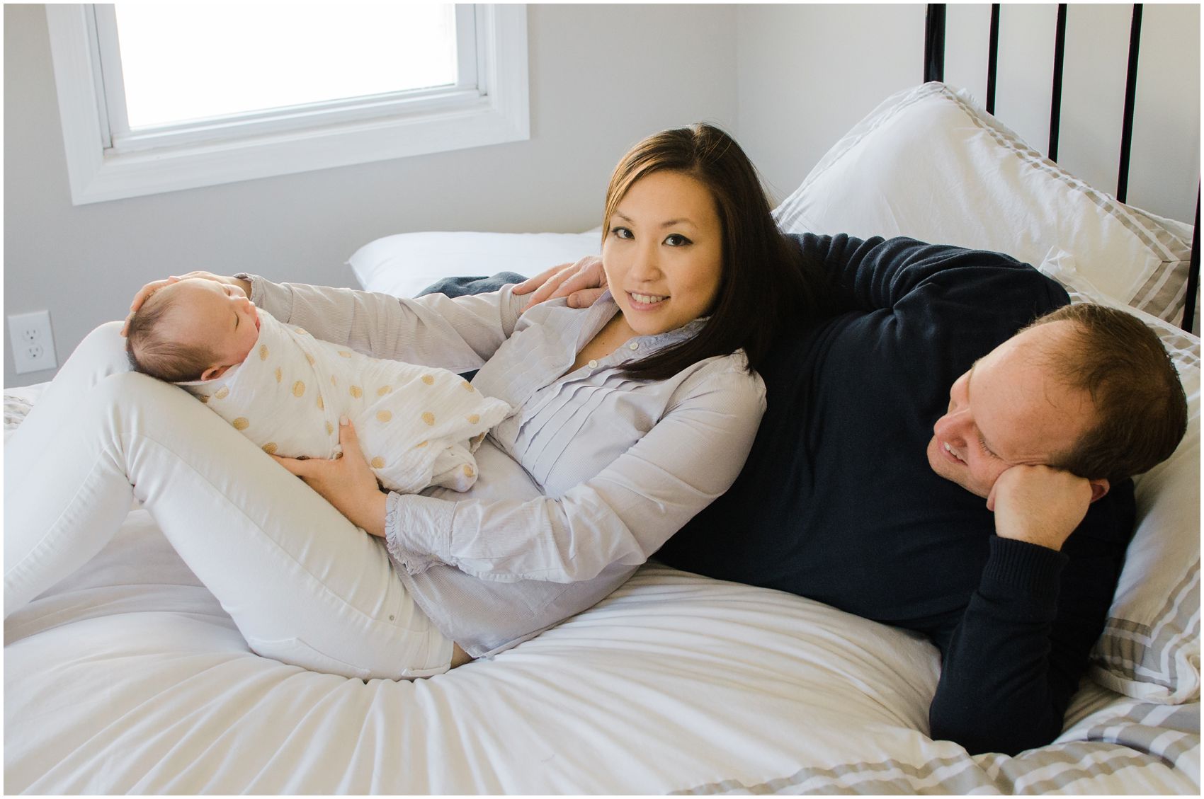 mom and dad laying on white bed with their new baby girl in NYC
