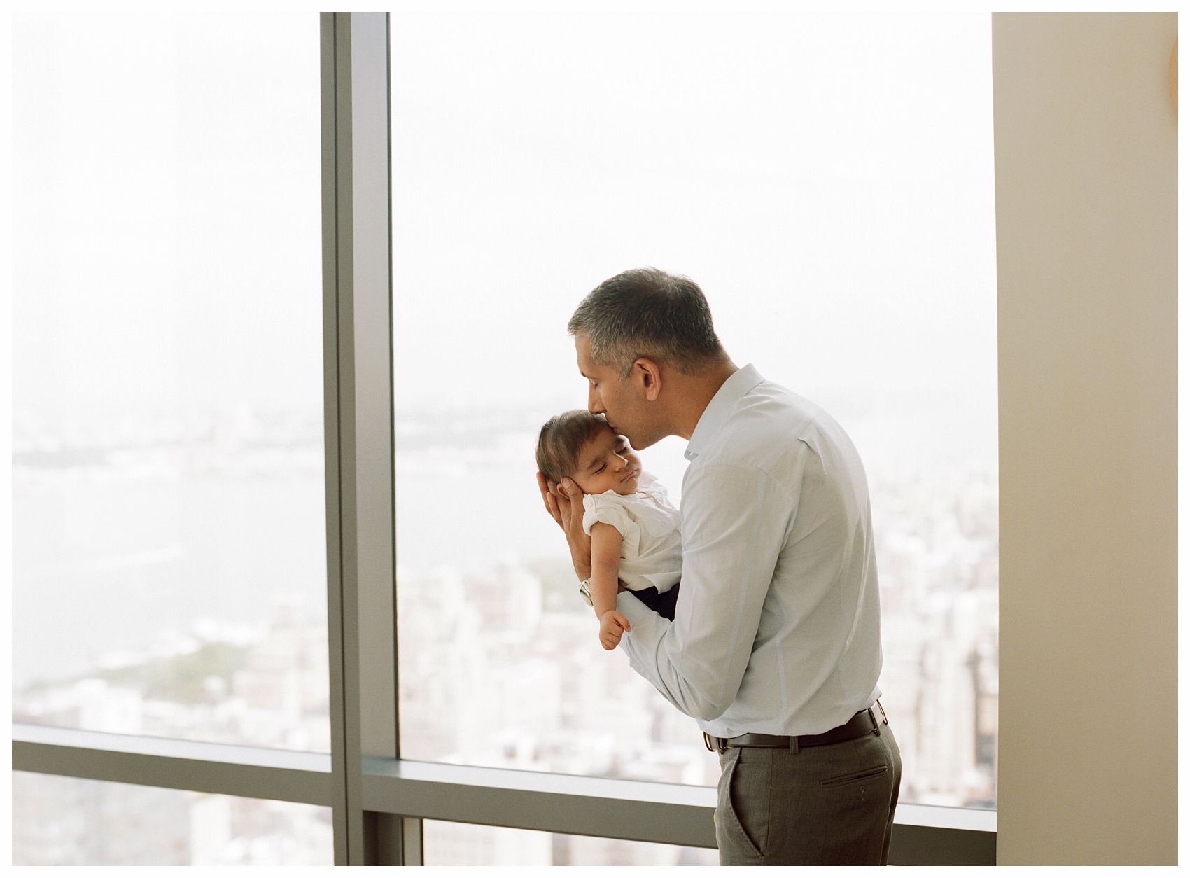 Best Newborn Photographer captures intimate moment of a father kissing his baby son lovingly by the big window overlooking NYC