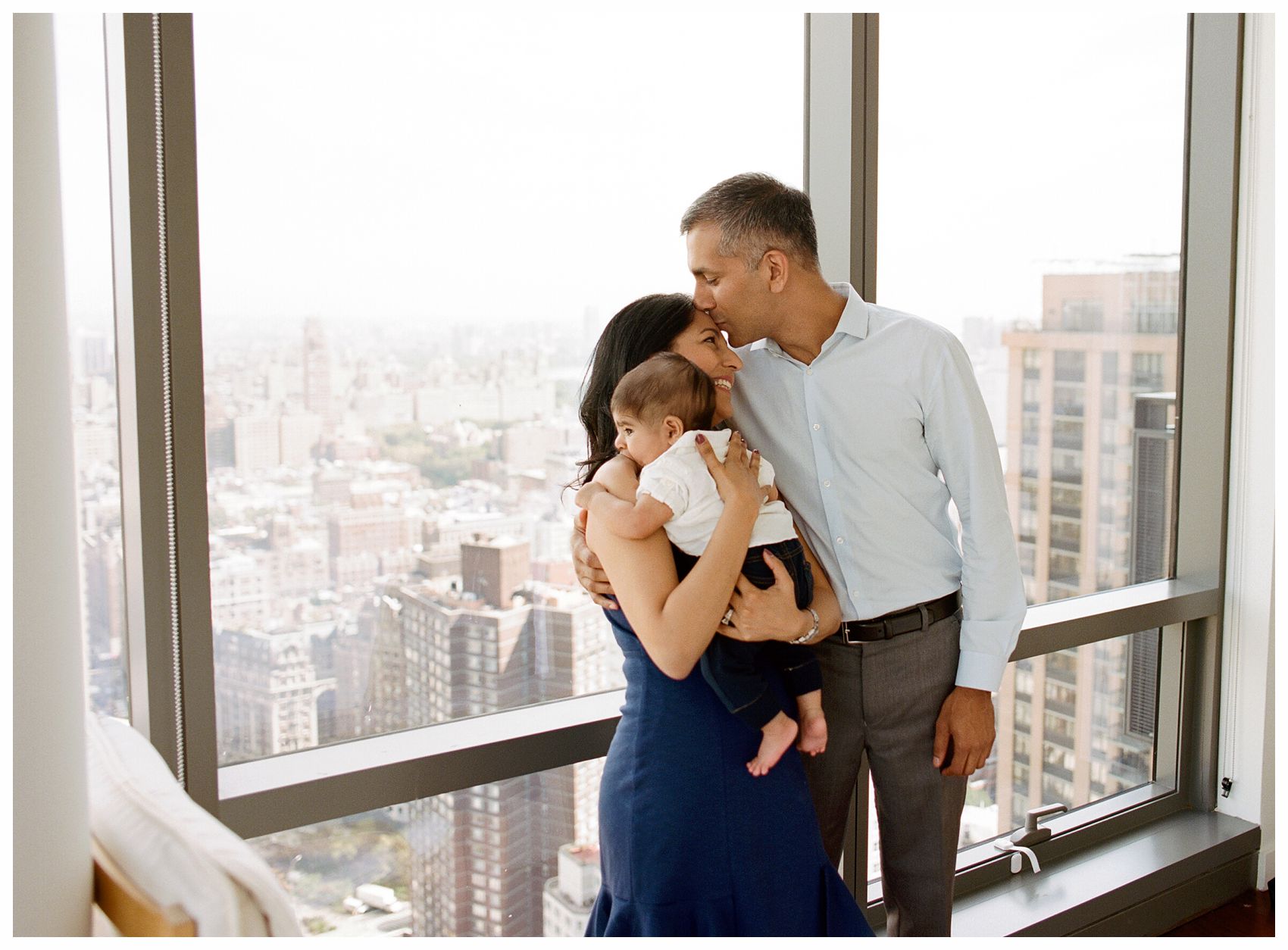 handsome dad kissing gorgeous mom and baby boy by the window in NYC during family photos