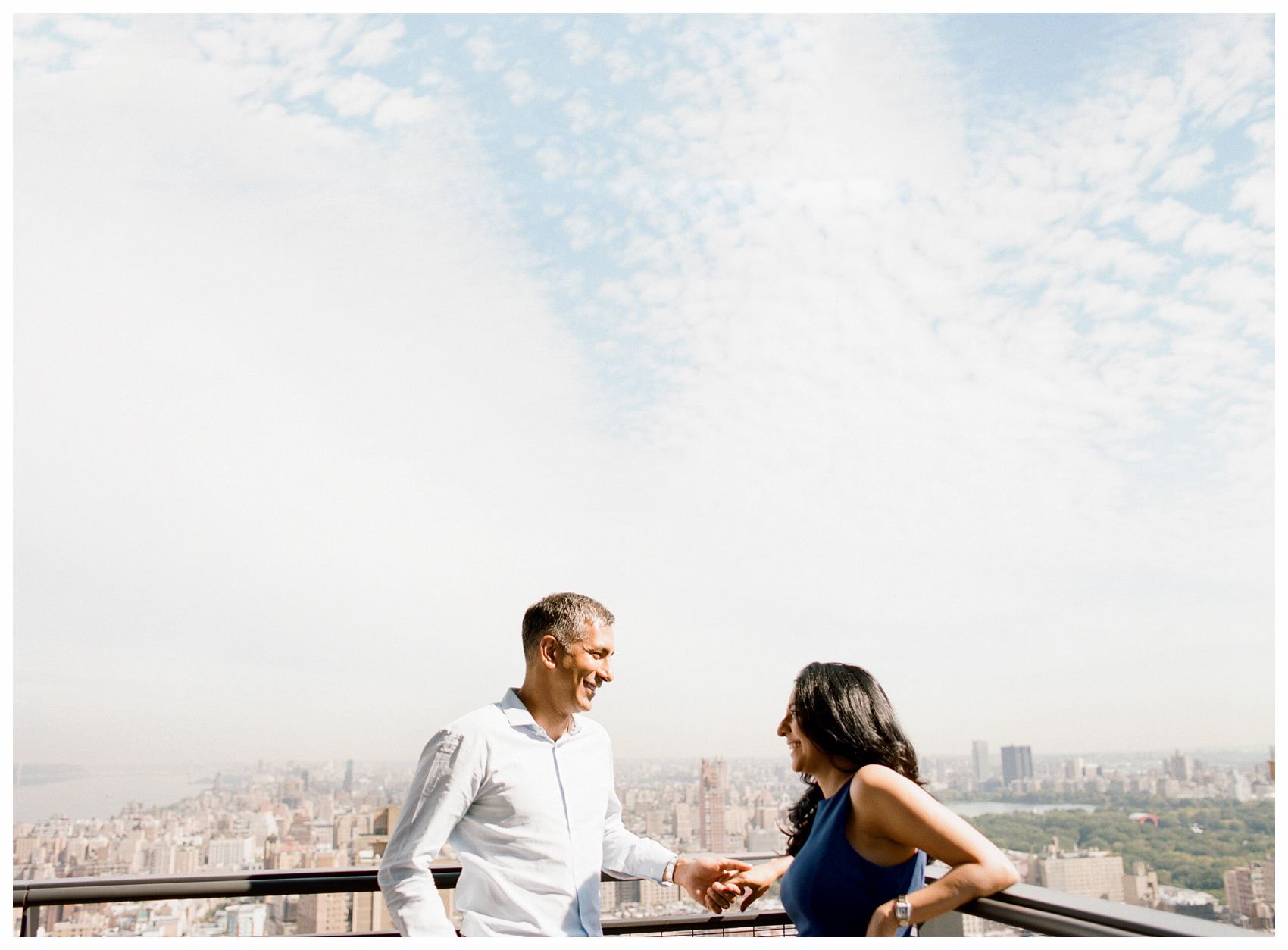 mom and dad holding hands and sharing an intimate happy moment against the NYC Skyline and the blue sky