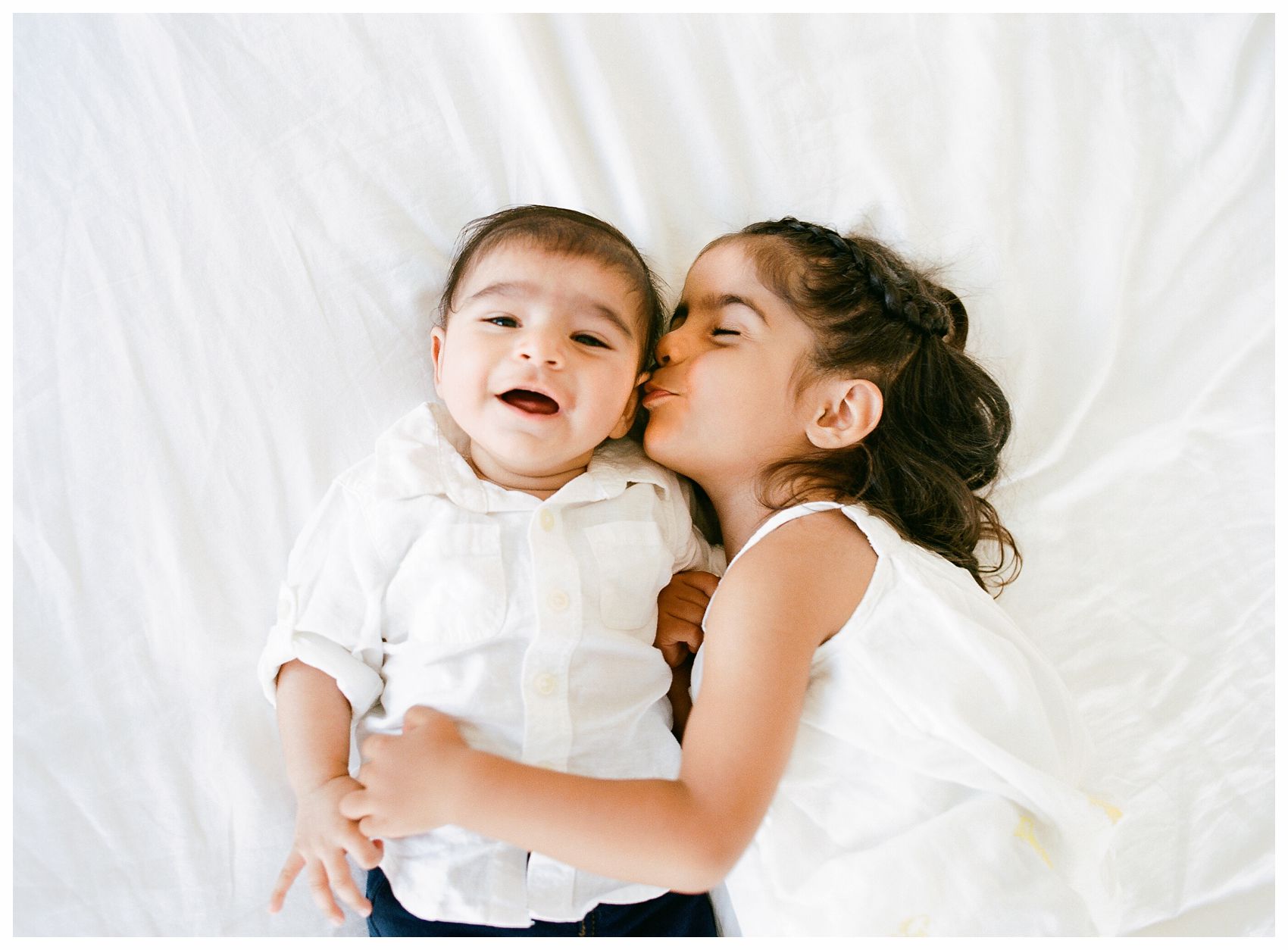 NYC family photography includes little sister kissing her happy baby brother on a big white bed