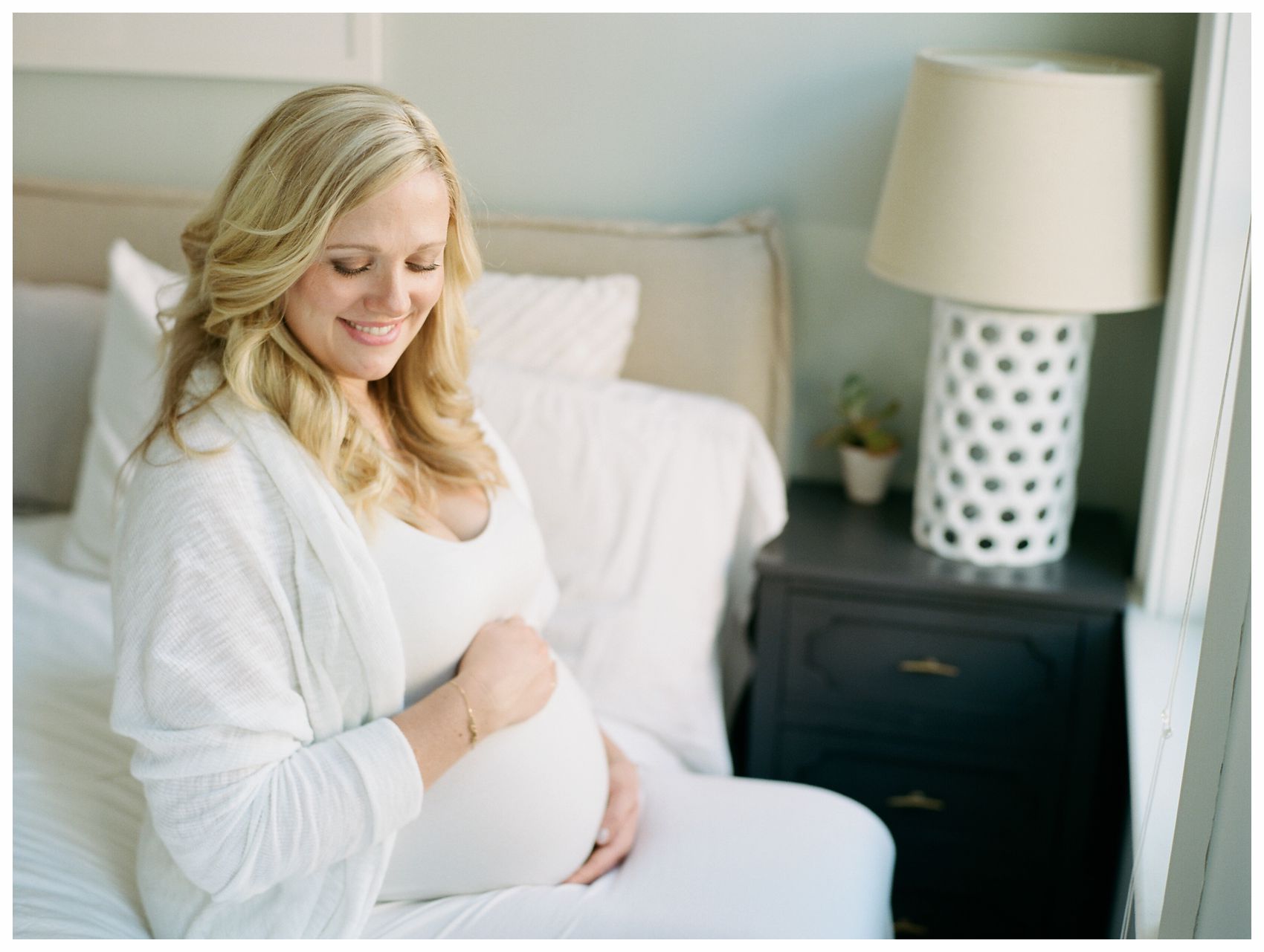 pregnant mom wearing a beautiful white dress and smiling at her belly in the in-home maternity session in Hoboken NJ
