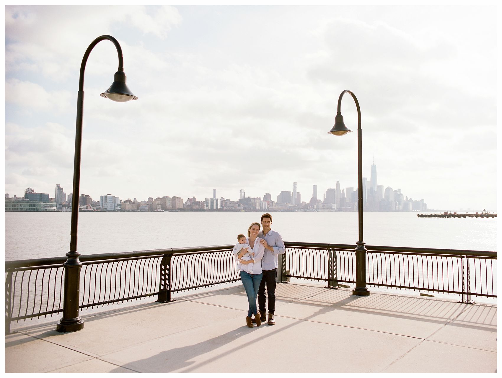 parents and the baby stand by NYC skyline at the Hoboken Waterfront 
